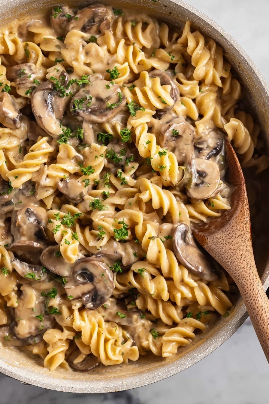 vegan mushroom stroganoff - The image shows a close-up of a white deep pan filled with creamy mushroom pasta. The pasta is spiral-shaped and has a yellowish color, mixed well with sliced light brown mushrooms. The sauce is thick and creamy, coating the pasta and mushrooms evenly. Small pieces of green parsley are sprinkled on top, adding specks of color. A wooden spoon is resting in the pan on the right side, partially submerged in the pasta. The pan is placed on a white marbled surface. photo taken with an iphone --ar 2:3 --v 7