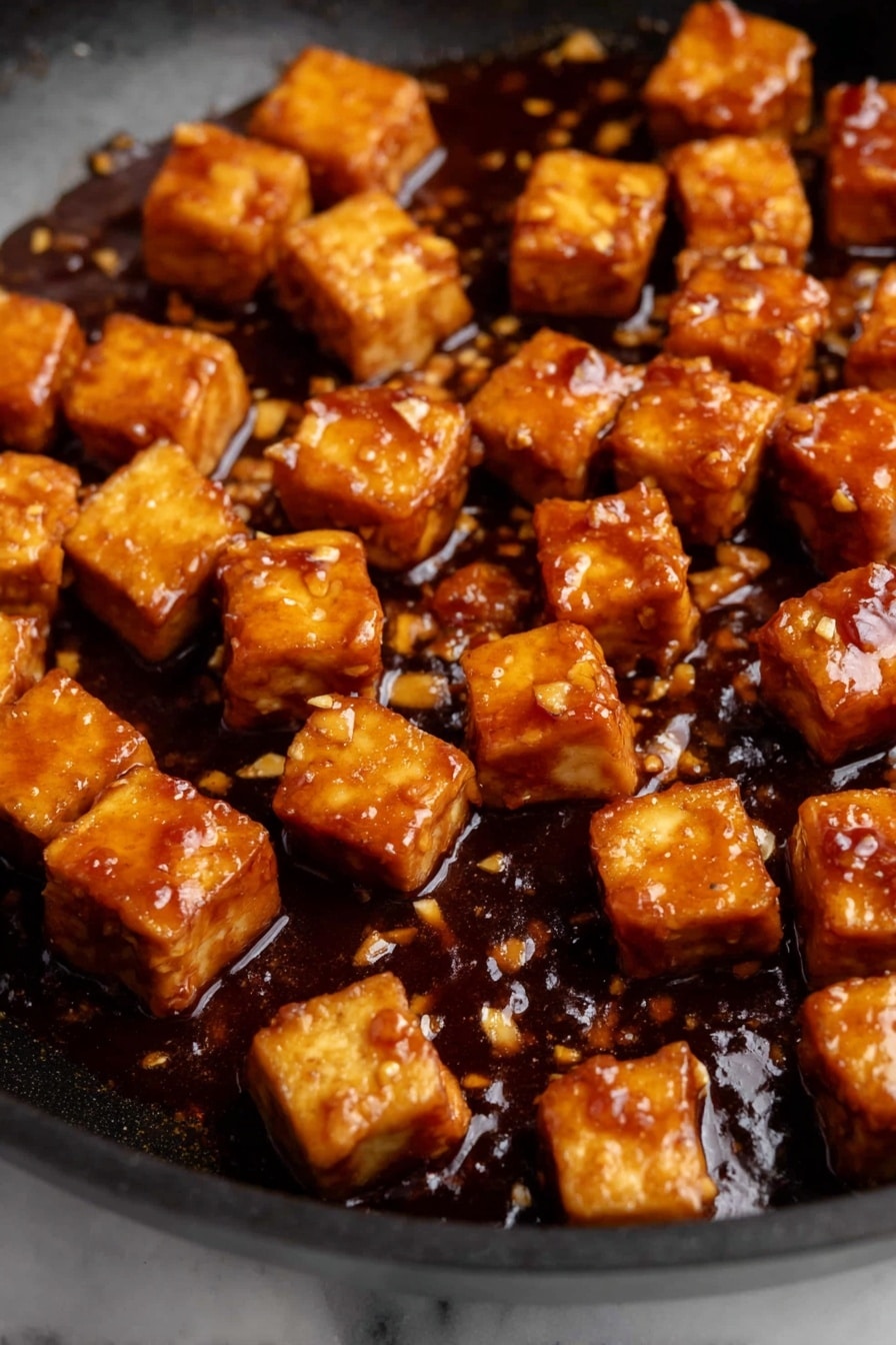Crispy Orange Tofu - The image shows many small, golden brown tofu cubes in a black pan, covered with a thick, shiny, dark brown sauce with small bits of garlic or seasoning scattered in it. The tofu cubes have a slightly crispy texture on the outside and are evenly coated with the sauce, which pools a little at the bottom of the pan. The background is a white marbled surface. photo taken with an iphone --ar 2:3 --v 7