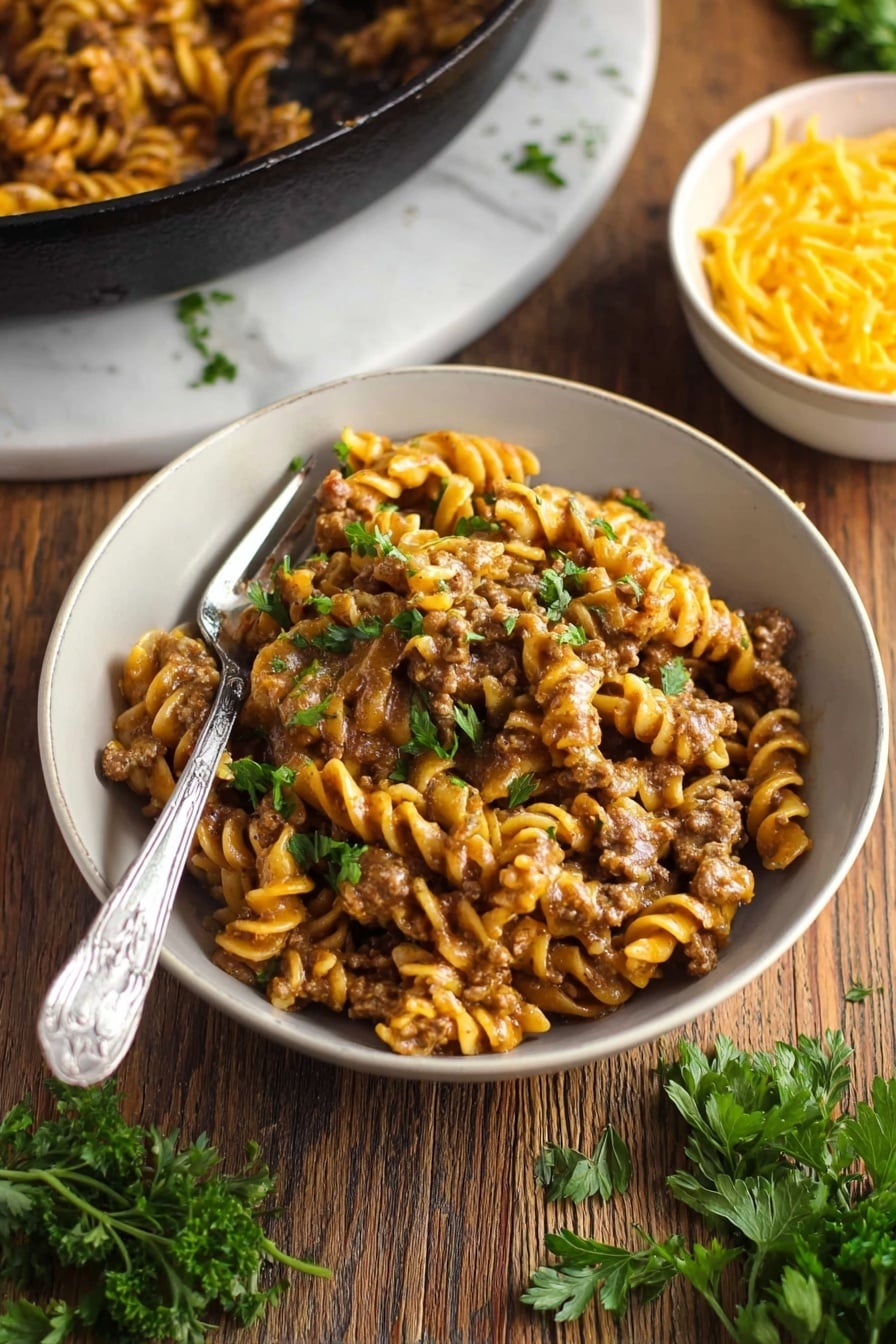 Vegan Cheeseburger Pasta - A black cast iron pan holds a creamy pasta dish with three layers: the bottom has a sauce mixed with ground plant-based meat that is dark brown and slightly chunky, the middle layer features short, spiral pasta in a light yellow-orange color coated in sauce, and the top layer is sprinkling of small green parsley pieces for garnish. The pan is placed on a black and white striped cloth on a wooden surface with uncooked spiral pasta scattered around. To the left of the pan, a white bowl contains shredded yellow cheddar cheese, and above is a package of plant-based meat. The background is a white marbled texture. Photo taken with an iphone --ar 2:3 --v 7