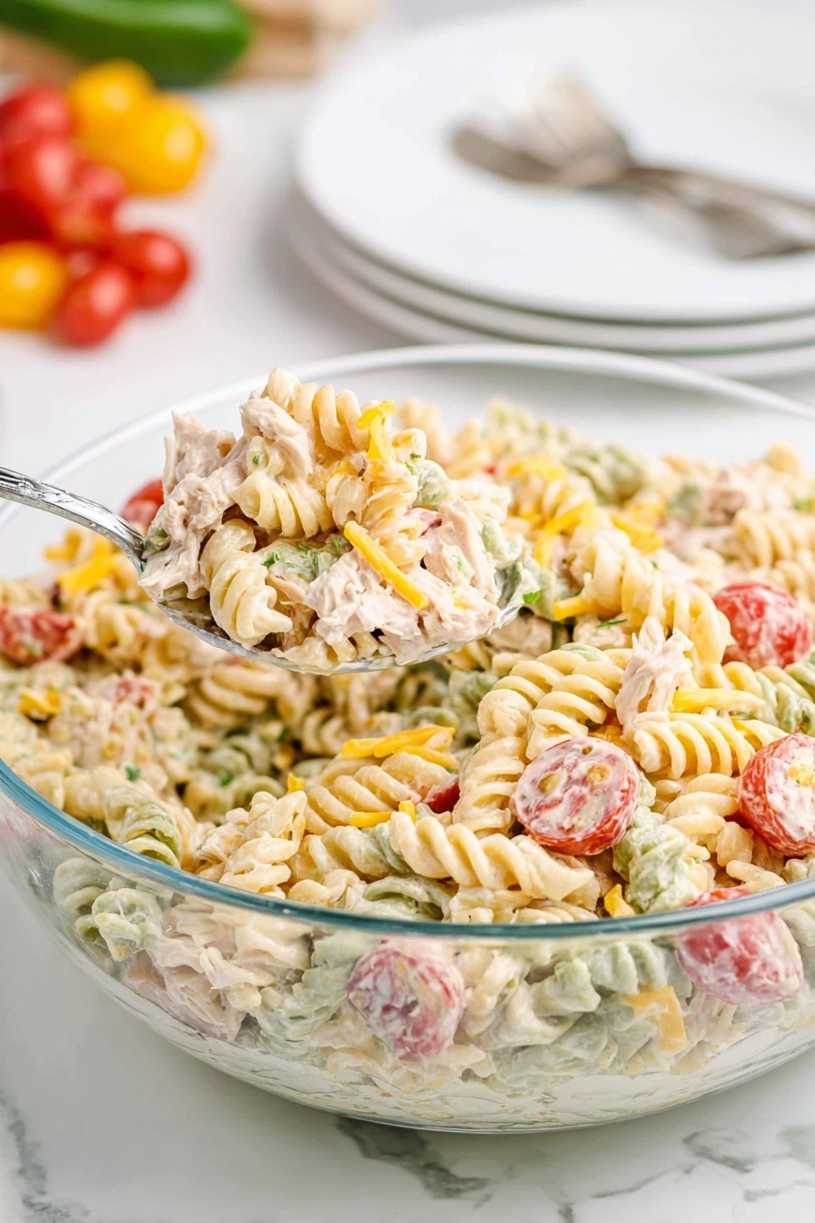 A clear glass bowl filled with tri-color spiral pasta coated in a creamy dressing sits on a white marbled surface. The pasta colors include a pale yellow, light green, and soft orange. Mixed within the pasta are halved red cherry tomatoes, small pieces of light beige chicken, and small bits of shredded yellow cheese scattered lightly on top. A metal spoon is scooping a portion from the bowl, showing the thick texture of the salad. In the blurred background, white plates with a fork and knife rest stacked, along with some whole cherry tomatoes and a green vegetable. Photo taken with an iphone --ar 2:3 --v 7