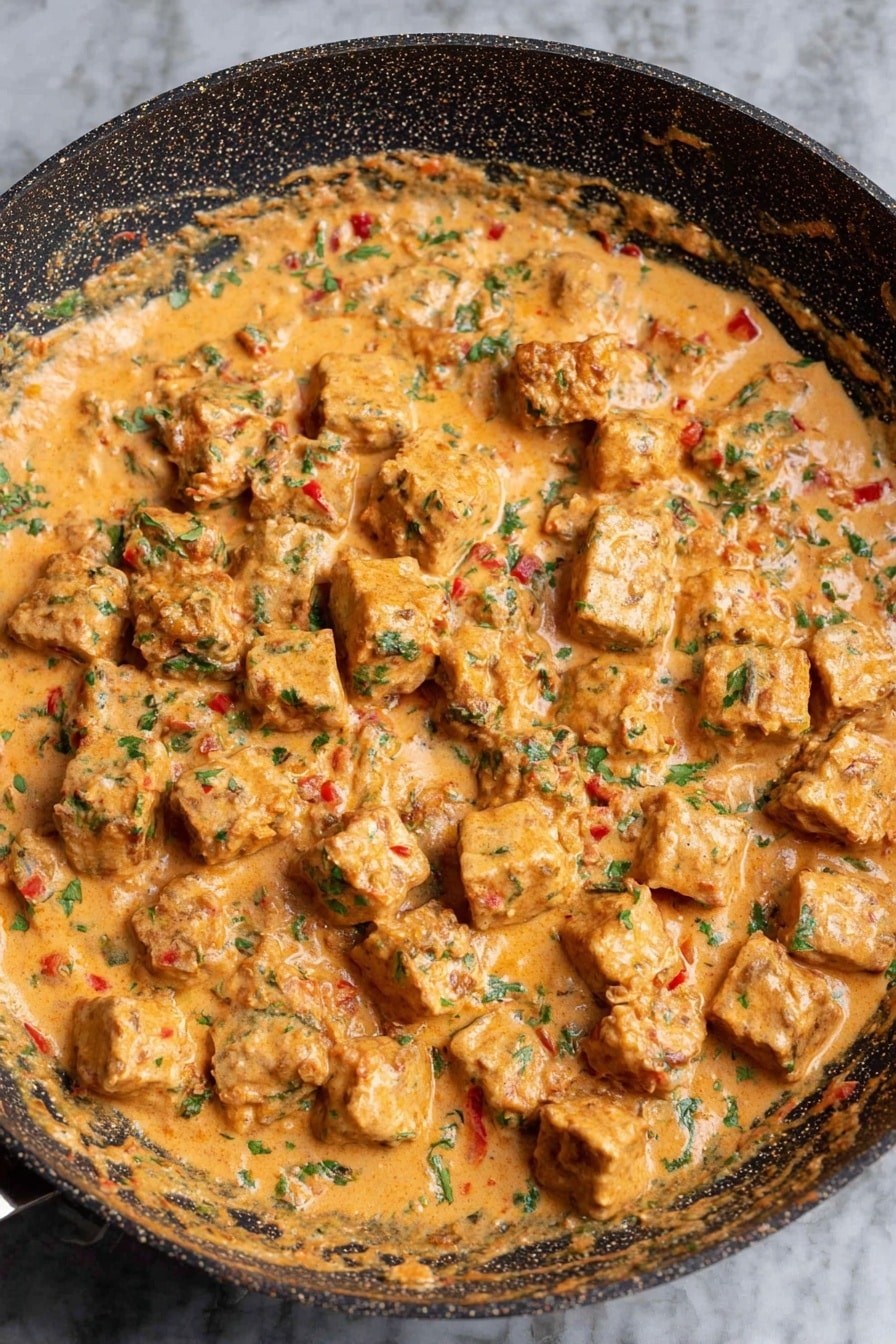 Marry Me Tofu - The image shows a pan filled with a creamy sauce that has a light orange color mixed with green herbs and small red bits. In the sauce, there are many evenly sized, light brown cubes that are partly covered by the sauce. The pan is round with a dark, patterned surface, and it sits on a white marbled background. The texture of the sauce looks thick and smooth with visible small pieces blending in photo taken with an iphone --ar 2:3 --v 7