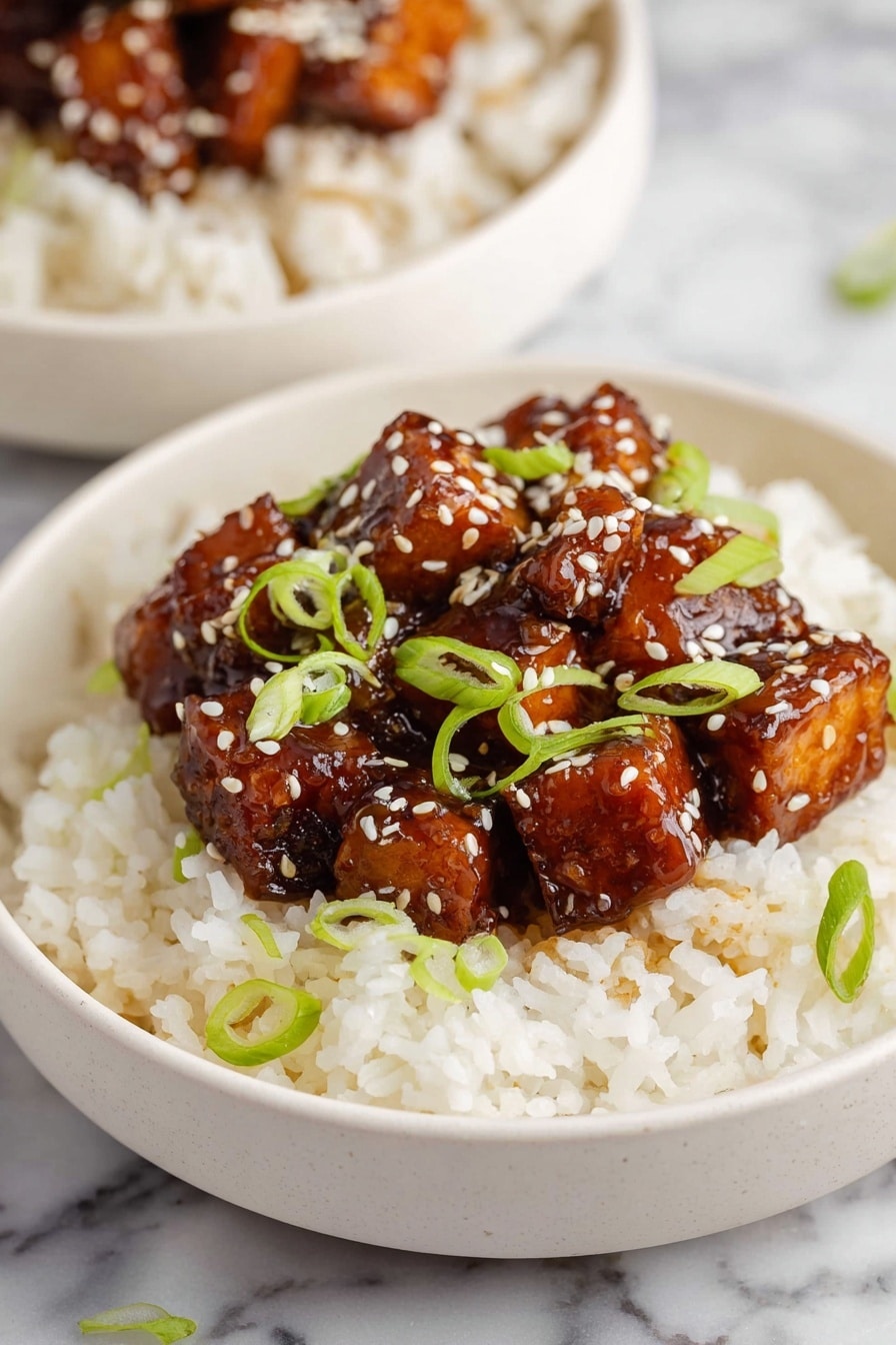 Crispy Orange Tofu - A white bowl filled with a base layer of fluffy white rice, with a thick layer of glossy, dark brown glazed tofu cubes placed in the center on top. The tofu pieces are sprinkled with white sesame seeds and decorated with thinly sliced green spring onions scattered across both the tofu and the rice. The bowl sits on a white marbled surface with another similar bowl blurred in the background. Photo taken with an iphone --ar 2:3 --v 7