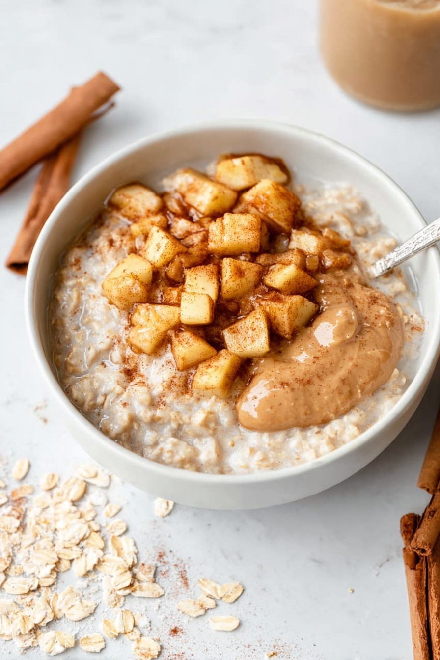 The image shows a white bowl filled with thick oatmeal topped with a generous layer of small golden-brown cooked apple cubes sprinkled with cinnamon, creating a textured and warm-colored top layer. The oatmeal beneath is creamy and light beige, with some visible grains giving it a slightly rough texture. A spoon scoops into the oatmeal, lifting a creamy portion mixed with the apple cubes. The bowl sits on a white marbled surface scattered lightly with oat flakes. Nearby, a woman's hand is holding the spoon from the right side. Around this bowl, there are glimpses of two more white bowls of oatmeal, one topped similarly with cooked apple cubes and the other with a swirl of smooth beige almond butter and cooked apples. There are also cinnamon sticks on the surface near the bowls. Photo taken with an iphone --ar 2:3 --v 7