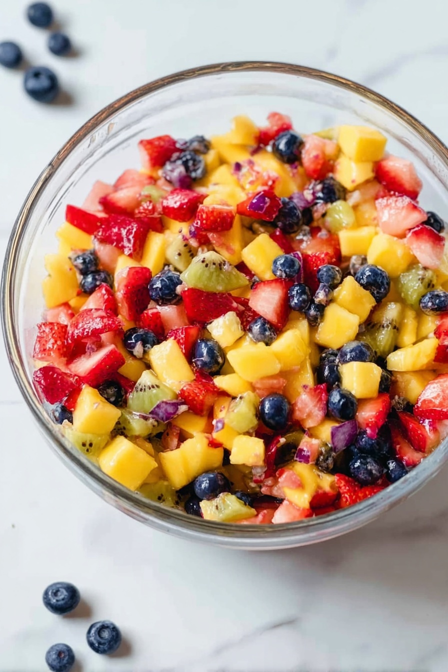 A clear glass bowl filled with a colorful fruit salad showing three main layers of mixed diced fruits: bright yellow mango chunks, red strawberry pieces, and dark blue whole blueberries, along with small bits of translucent kiwi adding texture. The fruits are mixed evenly, creating a vibrant patchwork of colors with a juicy, fresh appearance. The bowl sits on a white marbled surface with a few scattered blueberries nearby. The lighting highlights the natural shine and fresh look of the fruit. Photo taken with an iphone --ar 2:3 --v 7