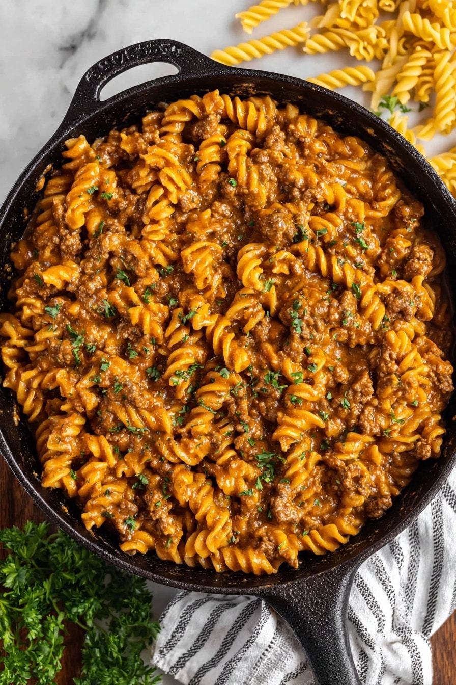 Vegan Cheeseburger Pasta - The image shows a black cast iron pan filled with creamy pasta. The pasta is spiral-shaped and covered in a thick orange-brown sauce mixed with pieces of cooked ground meat. Small green parsley leaves are scattered on top, adding contrast to the warm color of the sauce. The pan rests on a white striped cloth on a white marbled surface with some uncooked spiral pasta and parsley visible around it. Photo taken with an iphone --ar 2:3 --v 7