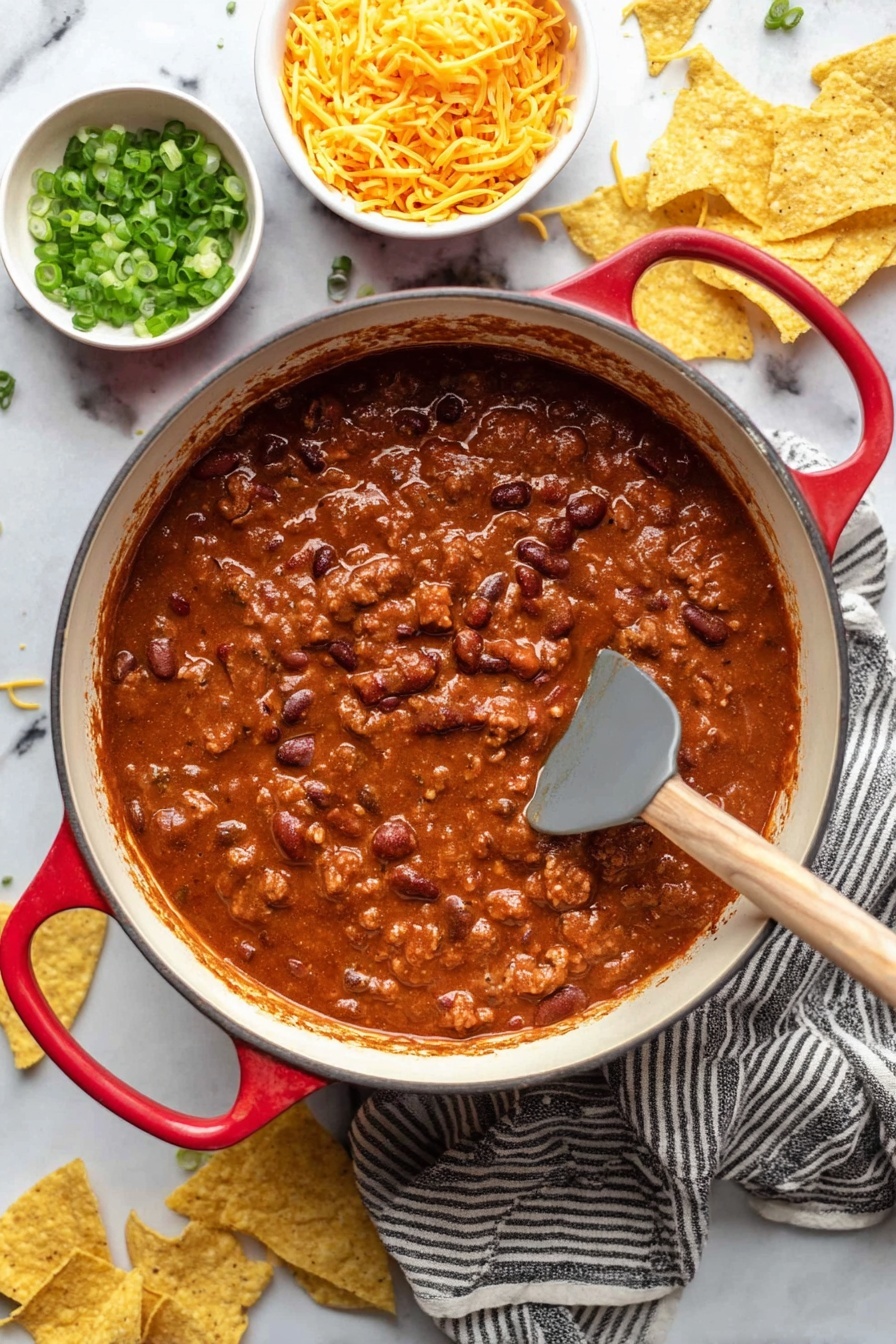 A large white pot with red handles is filled with thick, rich chili, showing beans and pieces of meat in a deep reddish-brown sauce, with a gray spatula with a wooden handle resting inside. Around the pot, there are yellow tortilla chips scattered on a white marbled surface, along with two small white bowls filled with shredded yellow cheese and chopped green onions. A striped cloth is placed underneath the pot. Photo taken with an iphone --ar 2:3 --v 7