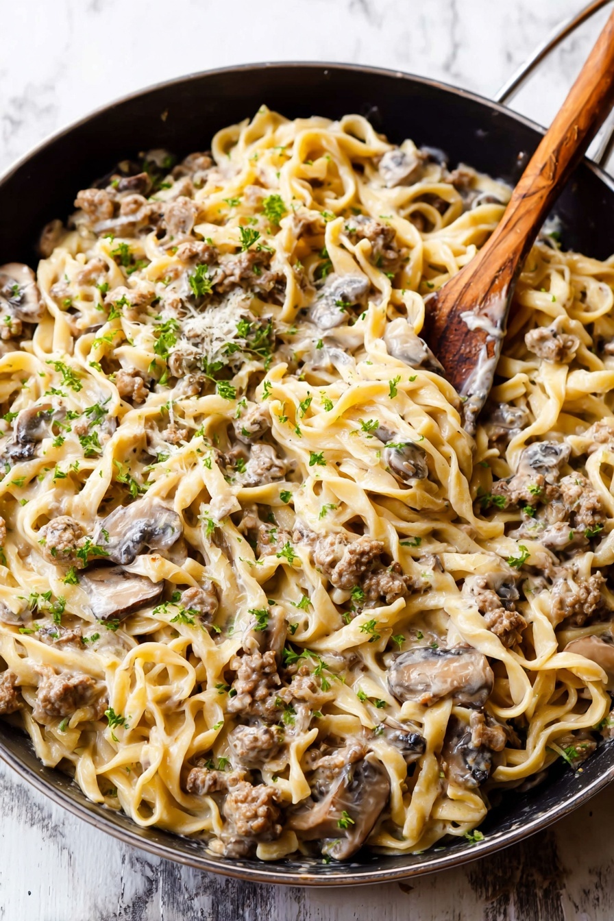 The dish is a creamy noodle mix served in a dark pan on a white marbled surface. There are about two layers: the first layer is twisted egg noodles that are light yellow with a soft, slightly chewy texture. The second layer consists of small browned mushrooms and crumbled cooked meat evenly mixed into the noodles. The whole dish is coated with a thick, light beige creamy sauce that holds everything together. Small green parsley pieces are sprinkled on top to add color. A wooden-handle spoon is in the pan, partly buried in the noodles. Photo taken with an iphone --ar 2:3 --v 7