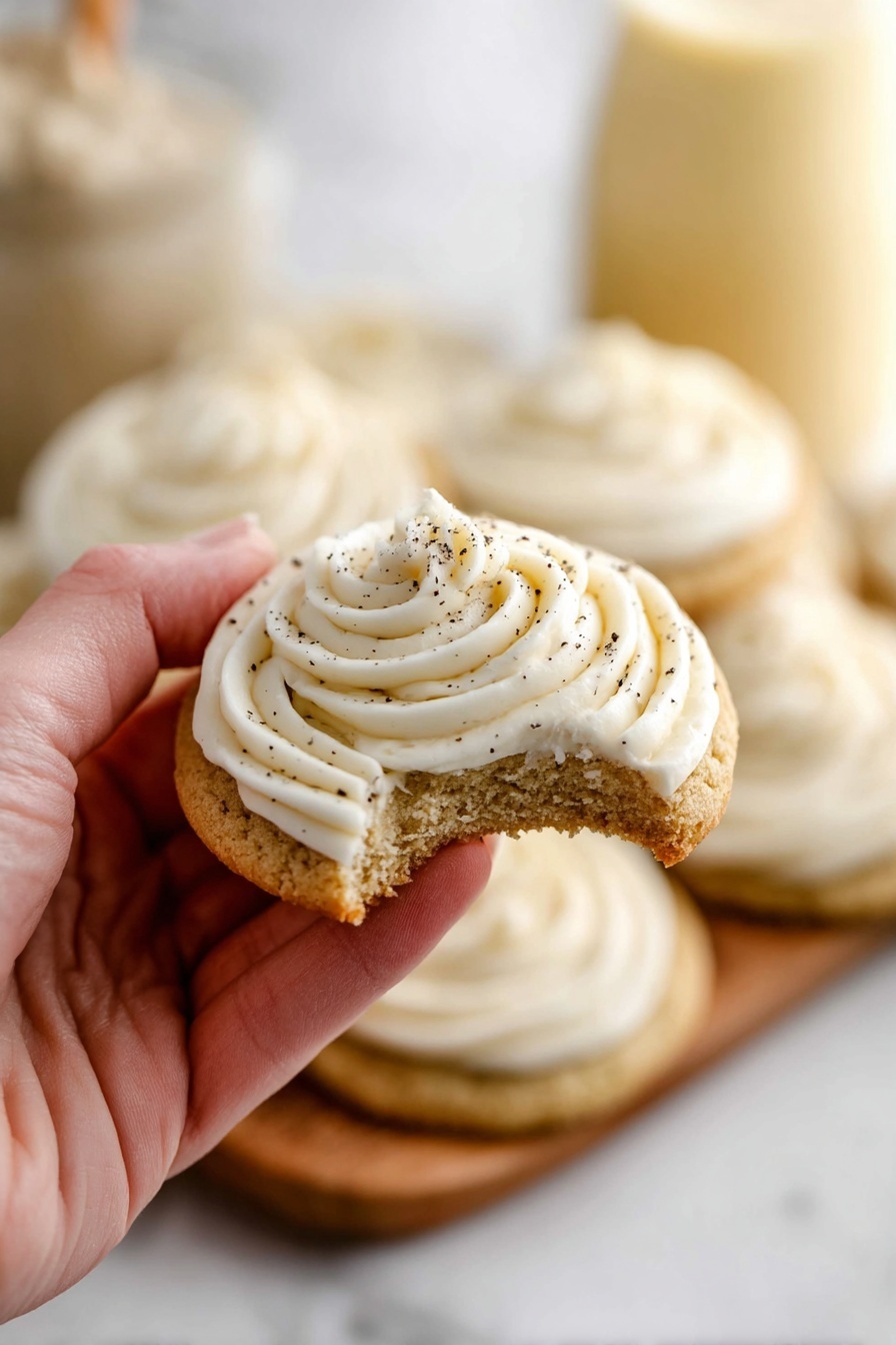 A close-up shows a woman's hand holding half of a soft light brown cookie with a visible crumb texture. On top of the cookie is a thick layer of smooth white frosting piped with swirls, sprinkled lightly with small dark specks. In the background, more cookies with the same frosting design sit on a white marbled surface, and blurred containers with light cream contents are visible. photo taken with an iphone --ar 2:3 --v 7
