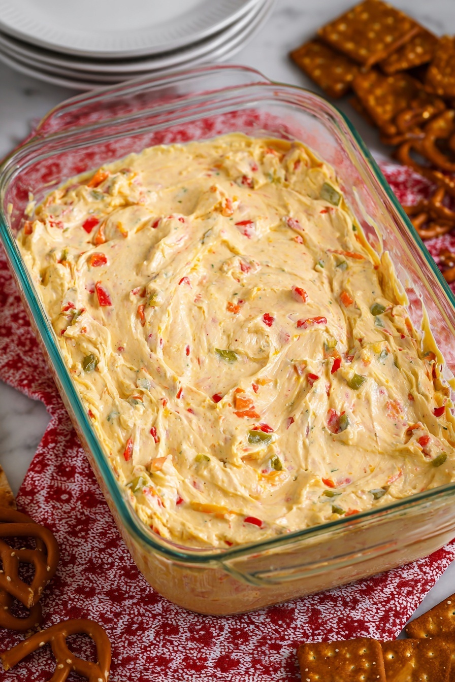 The image shows a rectangular glass dish filled with a creamy, spread-like mixture that has a soft, thick texture. The mixture is pale yellow with visible small pieces of red and green vegetables scattered evenly throughout. The surface of the mixture has some gentle swirls and slight peaks, giving it a smooth yet slightly textured look. The dish is placed on a red and white patterned cloth, and around the dish there are some brown pretzel snacks and a stack of white plates on a white marbled surface. Photo taken with an iphone --ar 2:3 --v 7