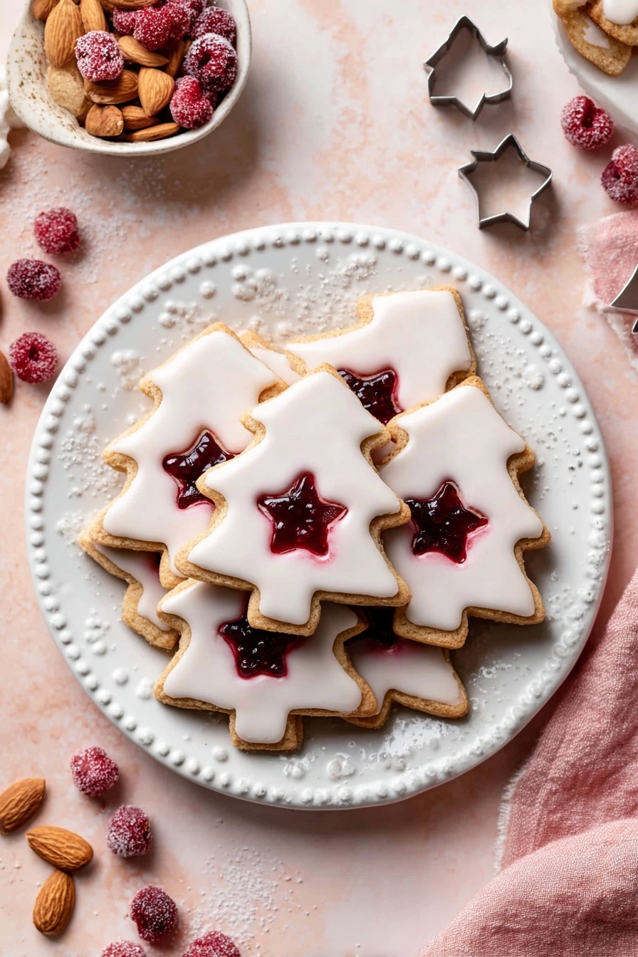 A white plate with a detailed edge holds a pile of tree-shaped cookies. Each cookie has two layers: the bottom layer is light brown and the top layer is covered with smooth white icing. The top layer has a star-shaped cutout in the center showing the red jam inside. Scattered around the plate are whole frozen red berries. The plate is on a pale pink surface with a white marbled texture just visible around the edges. A small white bowl with whole almonds and another with red berries sit nearby, along with some metal cookie cutters, and a pink cloth is partially seen. photo taken with an iphone --ar 2:3 --v 7