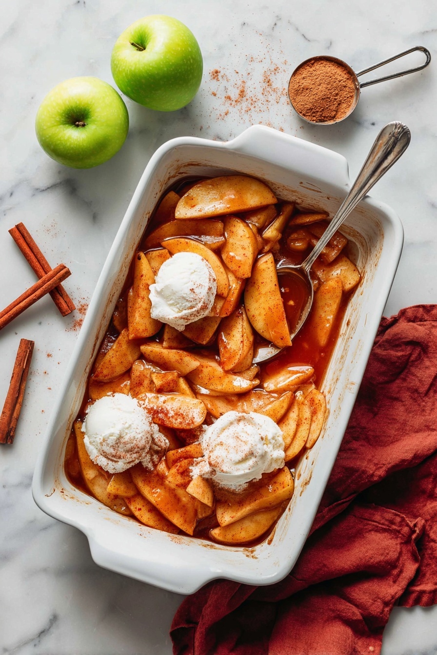 The image shows a white rectangular baking dish filled with warm, sliced apple pieces cooked in a rich, cinnamon-spiced sauce. The apple slices are golden brown and soft, piled unevenly with some sauce pooling at the bottom. There are two small scoops of creamy white vanilla ice cream placed on top of the apples, one near the edge and one near the center. A silver spoon rests inside the dish, partially buried in the apples. Around the dish, on a white marbled surface, there are cinnamon sticks, a measuring cup with brown sugar, a few fresh green apples, and a reddish cloth napkin partially tucked under the dish. Photo taken with an iphone --ar 2:3 --v 7
