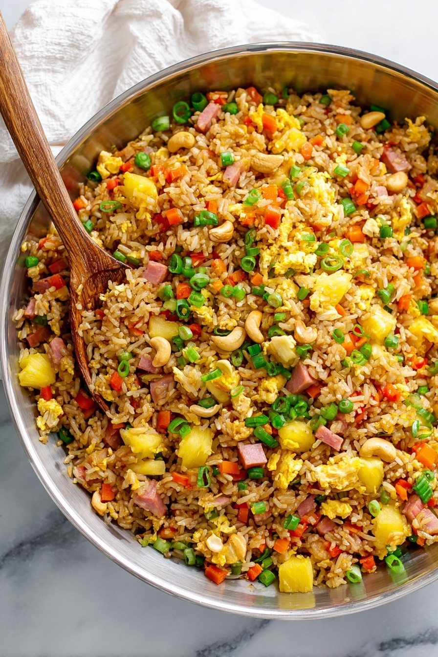 A large silver bowl filled with fried rice showing many mixed layers: the base is brown fried rice with visible grains, mixed with small chunks of yellow scrambled egg scattered throughout, bright yellow pieces of pineapple, small diced pink ham, finely chopped orange carrot bits, diced red bell pepper, sliced green onions sprinkled on top, and whole light brown cashew nuts adding texture. A silver spoon rests inside the bowl, slightly digging into the rice. The bowl sits on a white marbled surface with a white cloth partly visible in the background. Photo taken with an iphone --ar 2:3 --v 7
