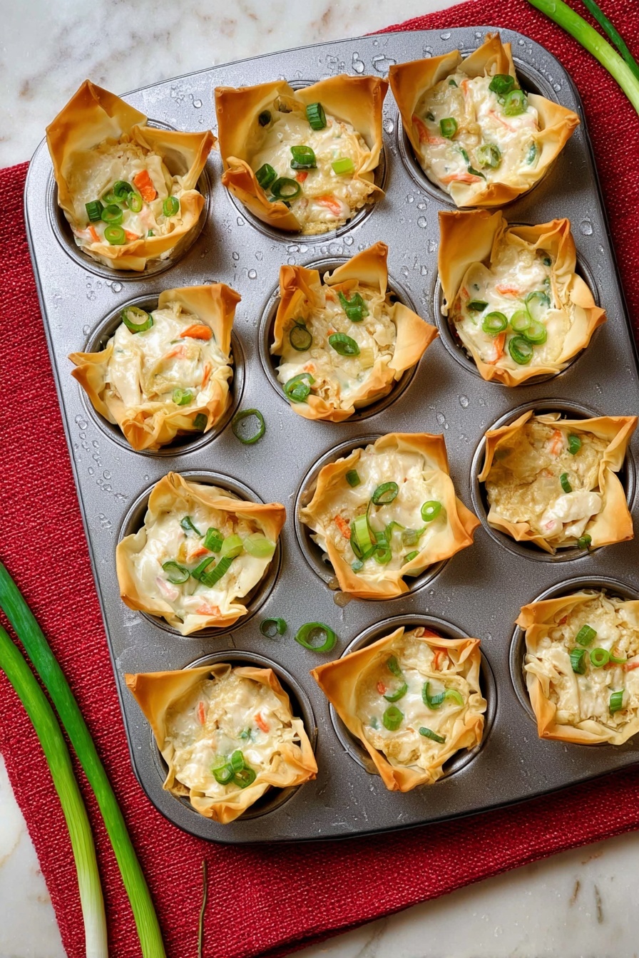 A metal muffin tray holds twelve baked cups made from golden brown, crispy wonton wrappers. Each cup is filled with a creamy white mixture dotted with small pieces of orange carrot and green scallion slices. Some scallion slices are also sprinkled on top of the mixture and around the cups on the tray. The tray sits on a red textured cloth over a white marbled surface. Two green scallion stalks lay next to the tray. Moisture droplets are visible on the metal tray. photo taken with an iphone --ar 2:3 --v 7