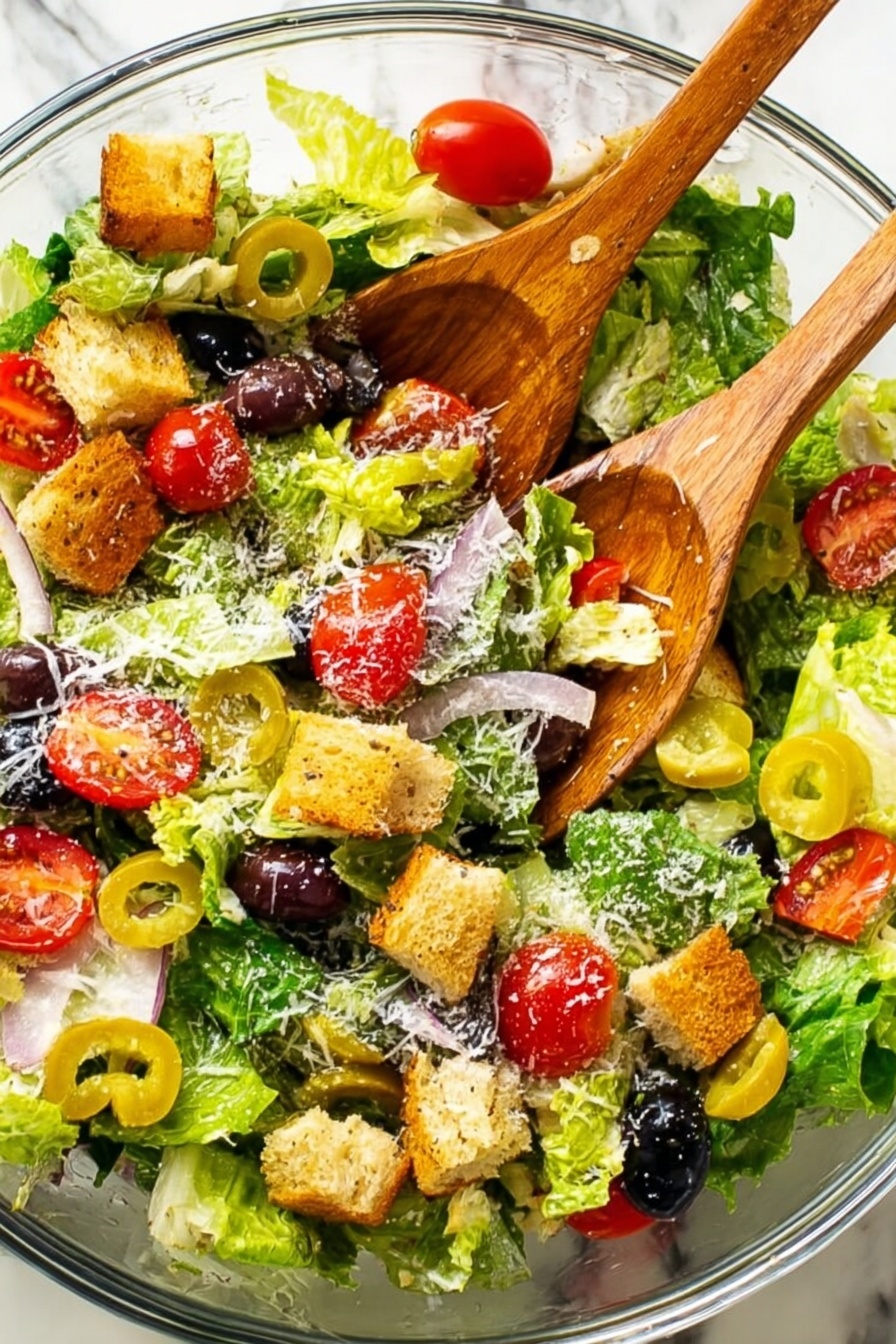 A clear glass bowl filled with a fresh salad showing multiple layers: the base is made of bright green leafy lettuce, topped with sliced red cherry tomatoes, whole black olives, and light yellow pepperoncini peppers. Scattered on top are golden brown croutons and a light sprinkling of grated cheese. Two wooden salad spoons rest inside the bowl on the salad. The bowl sits on a white marbled surface photo taken with an iphone --ar 2:3 --v 7