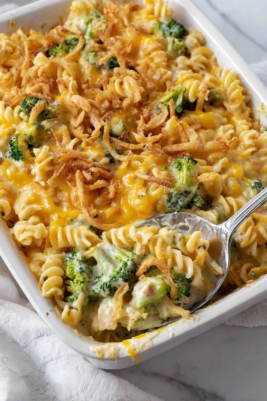 This image shows a white baking dish filled with a creamy pasta casserole. The pasta is rotini, with a mix of yellow cheddar cheese melted on top and creamy white sauce mixed throughout. There are bright green broccoli pieces scattered evenly in the dish. The top is sprinkled with light brown crispy fried onion rings, adding a crunchy texture. A silver spoon is scooping a portion, revealing layers of pasta, cheese, broccoli, and sauce. The background is a white marbled surface with a white cloth napkin beside the dish. Photo taken with an iphone --ar 2:3 --v 7
