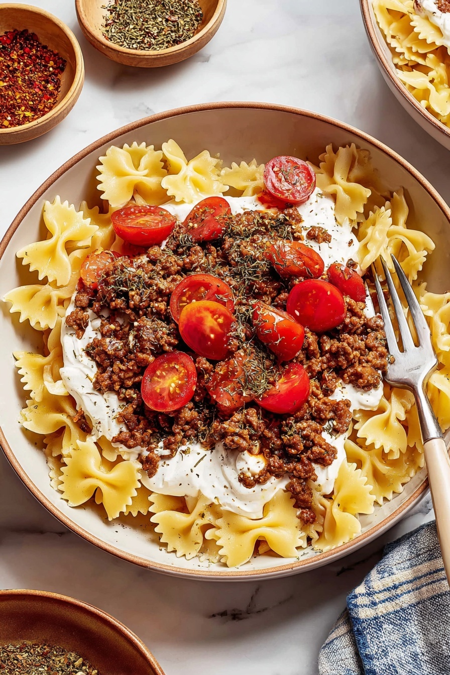 A white bowl filled with three clear layers: the bottom layer is light yellow bowtie pasta spread evenly, the middle layer is thick white sauce dolloped over the pasta, and the top layer is dark brown cooked ground meat mixed with spices and some small onion pieces, topped with halved bright red cherry tomatoes sprinkled with dried herbs. A fork is resting inside the bowl on the right side. The bowl is placed on a white marbled surface with a small beige bowl of ground spice and a gray bowl of white sauce nearby, along with another similar bowl of the same dish partially visible in the background. photo taken with an iphone --ar 2:3 --v 7