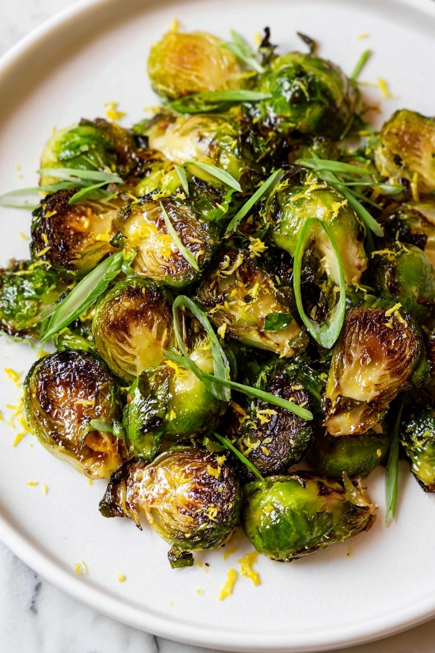 A white plate holds one layer of roasted Brussels sprouts, some showing golden brown and slightly charred edges while others are darker and more crisp. The Brussels sprouts are decorated with thin slices of bright green scallions scattered on top, and a light sprinkle of finely grated yellow zest adds texture and color contrast. The vegetables have a shiny glaze, suggesting a drizzle of sauce or oil. The plate rests on a white marbled surface. photo taken with an iphone --ar 2:3 --v 7