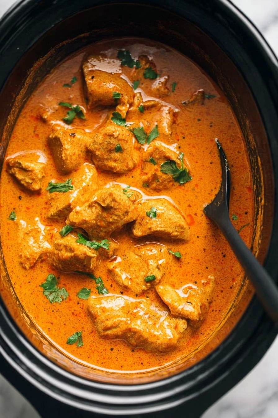 The image shows a close-up of a black slow cooker filled with a creamy orange curry sauce with several pieces of tender meat fully covered in the sauce. Small green herb pieces are sprinkled on top, adding a fresh touch. A black spoon is partially dipped into the curry on the right side, showing the thick and smooth texture of the sauce. The background is a white marbled surface. photo taken with an iphone --ar 2:3 --v 7